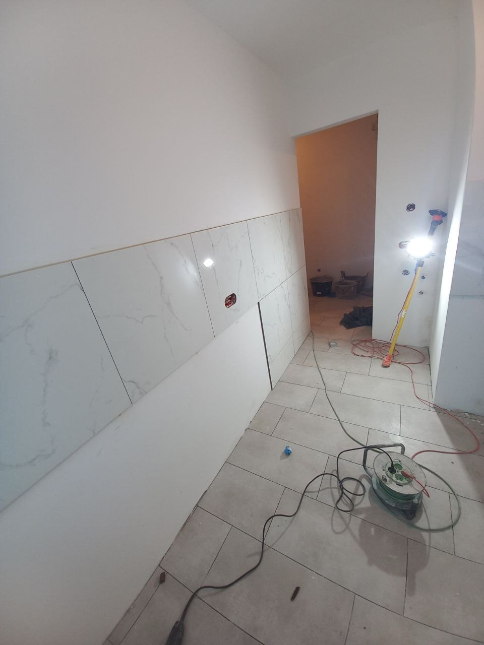 Wnętrze w trakcie prac wykończeniowych: częściowo wyłożona marmurowymi płytkami ściana, przewody elektryczne i zwinięty przedłużacz na podłodze z płytek, lampa robocza oświetlająca przestrzeń.