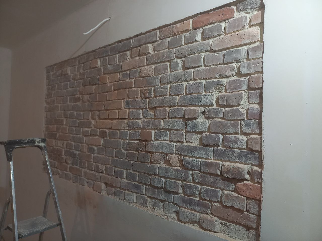 Fragment ściany wewnętrznej z odsłoniętą, starą cegłą, częściowo pokrytą szarą zaprawą, z widoczną drabiną malarską po lewej stronie.