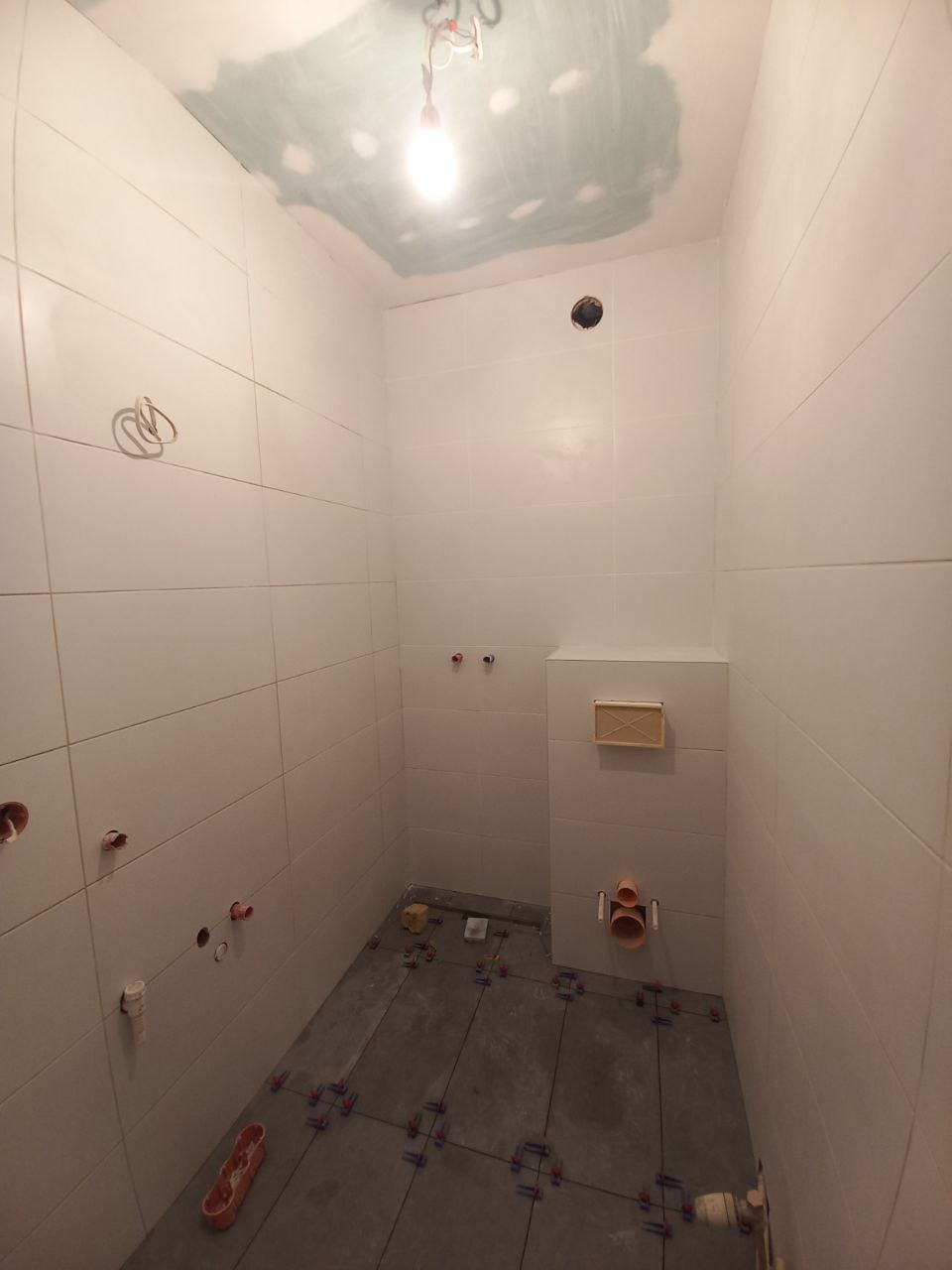 Wnętrze łazienki w trakcie remontu: białe płytki ścienne, szare płytki podłogowe w trakcie układania z użyciem krzyżyków dystansowych, widoczne instalacje wodne i elektryczne, sufit częściowo...