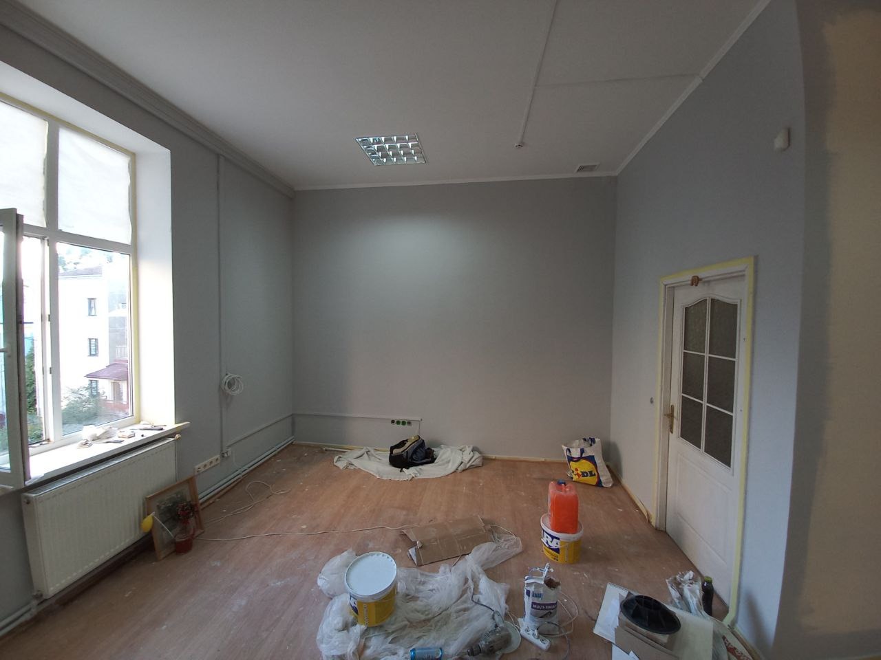 Pokój po świeżym pomalowaniu ścian na szaro, z widocznymi narzędziami malarskimi i zabezpieczeniami podłogi, drzwi oklejone taśmą malarską, widok z okna na budynek mieszkalny.