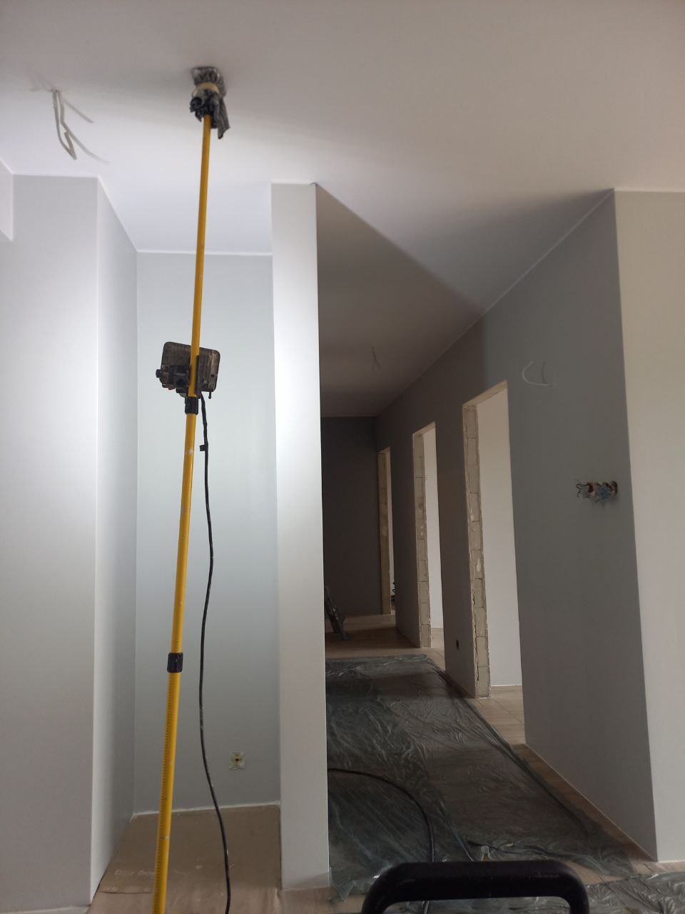 Wnętrze w trakcie odświeżania ścian i sufitu za pomocą wałka na teleskopowym kiju, podłoga zabezpieczona folią malarską, widoczne puszki elektryczne.