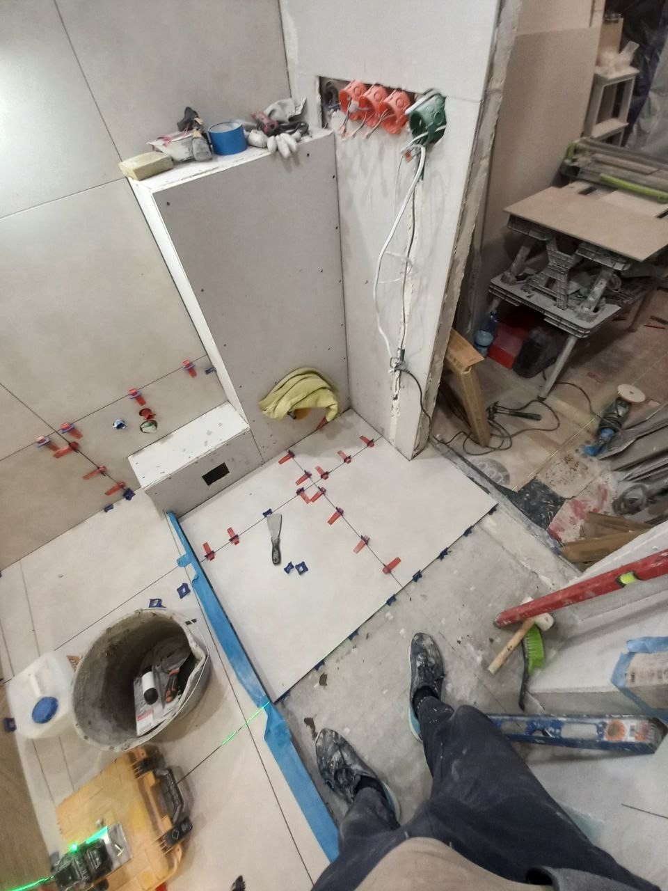 Układanie jasnych płytek podłogowych w łazience, widoczne krzyżyki dystansowe, poziomica laserowa, wiadro z zaprawą i narzędzia glazurnicze, instalacja elektryczna w ścianie, ujęcie z perspektywy...