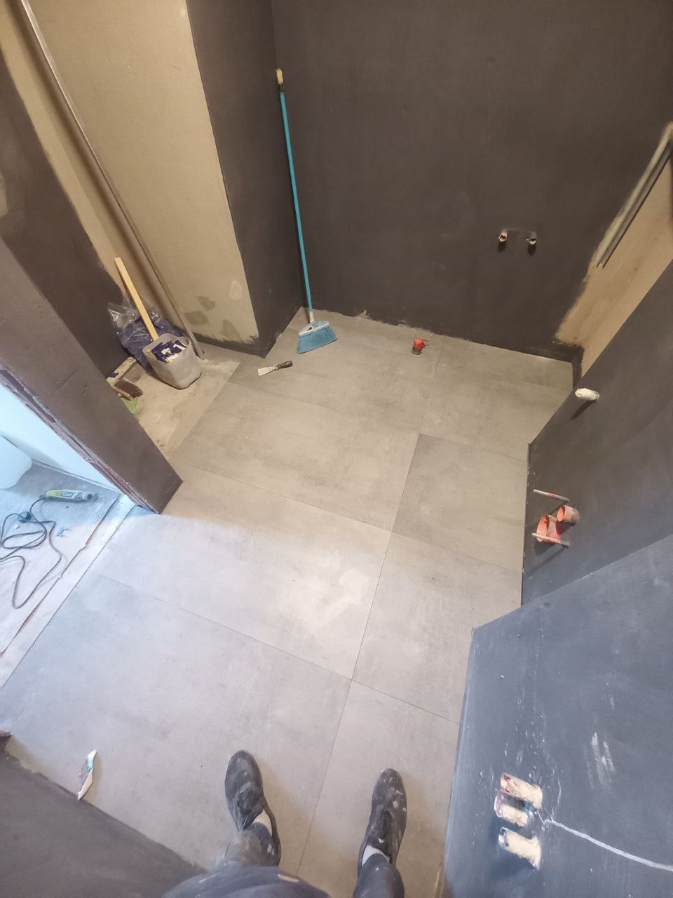 Świeżo ułożone, szare płytki podłogowe w łazience w trakcie remontu, widoczne narzędzia glazurnicze, ślady po szpachlowaniu ścian i fragment ubrań osoby robiącej zdjęcie.