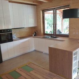 Andrzej Kozak OMKA-MEBLE - Nowoczesna kuchnia z białymi, lakierowanymi frontami szafek, drewnianymi ścianami i blatem, oraz czarną lodówką Samsung i dwiema czarnymi lampami wiszącymi; widoczny fragment układanej podłogi.