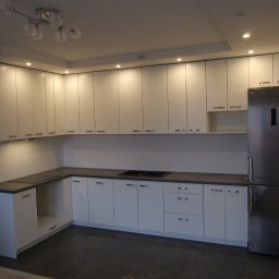 Andrzej Kozak OMKA-MEBLE - Nowoczesna, minimalistyczna kuchnia z białymi frontami szafek i ciemnoszarym blatem, oświetlona punktowymi lampami sufitowymi i wiszącą lampą z kilkoma szklanymi kloszami, z lodówką w folii ochronnej.