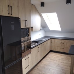 Andrzej Kozak OMKA-MEBLE - Jasna kuchnia w skandynawskim stylu na poddaszu ze skośnym sufitem i oknem dachowym, czarne blaty i sprzęt AGD, drewniana podłoga.