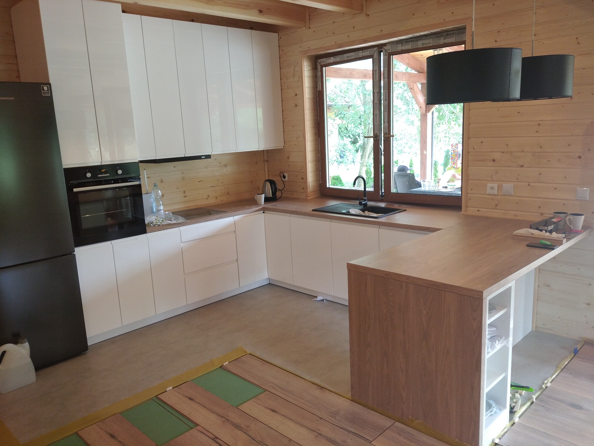 Nowoczesna kuchnia z białymi, lakierowanymi frontami szafek, drewnianymi ścianami i blatem, oraz czarną lodówką Samsung i dwiema czarnymi lampami wiszącymi; widoczny fragment układanej podłogi.