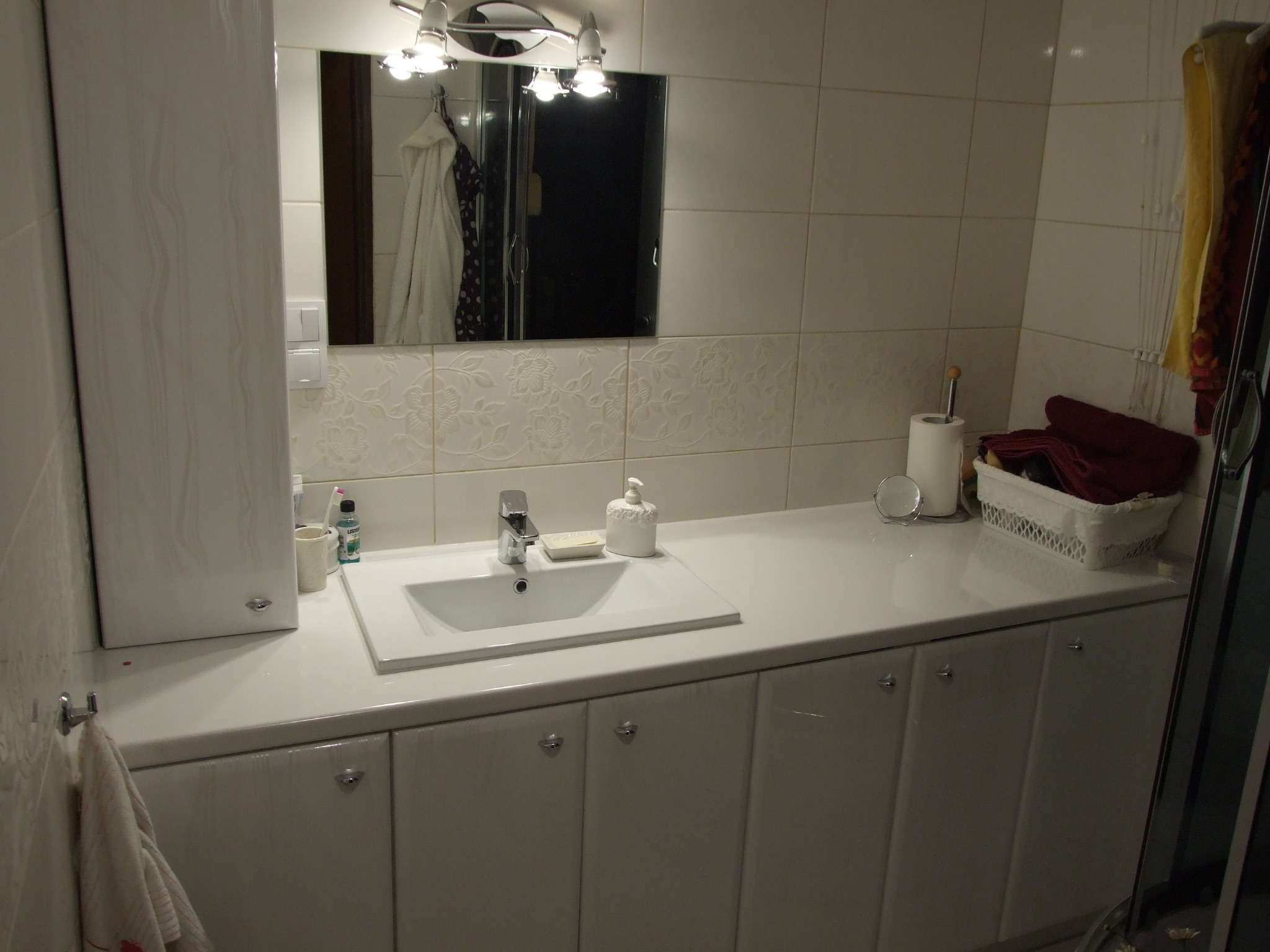 Biała łazienka z szafkami na wymiar pod umywalką, lustrem i ceramicznymi kaflami z kwiatowym wzorem na ścianie.