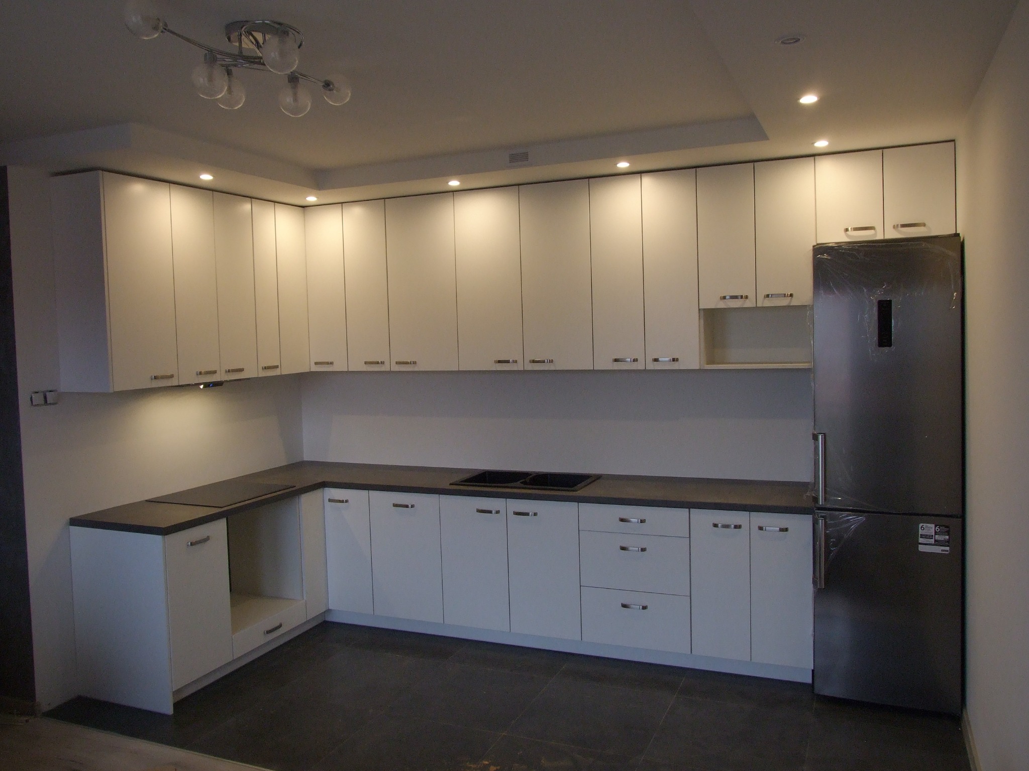 Nowoczesna, minimalistyczna kuchnia z białymi frontami szafek i ciemnoszarym blatem, oświetlona punktowymi lampami sufitowymi i wiszącą lampą z kilkoma szklanymi kloszami, z lodówką w folii ochronnej.