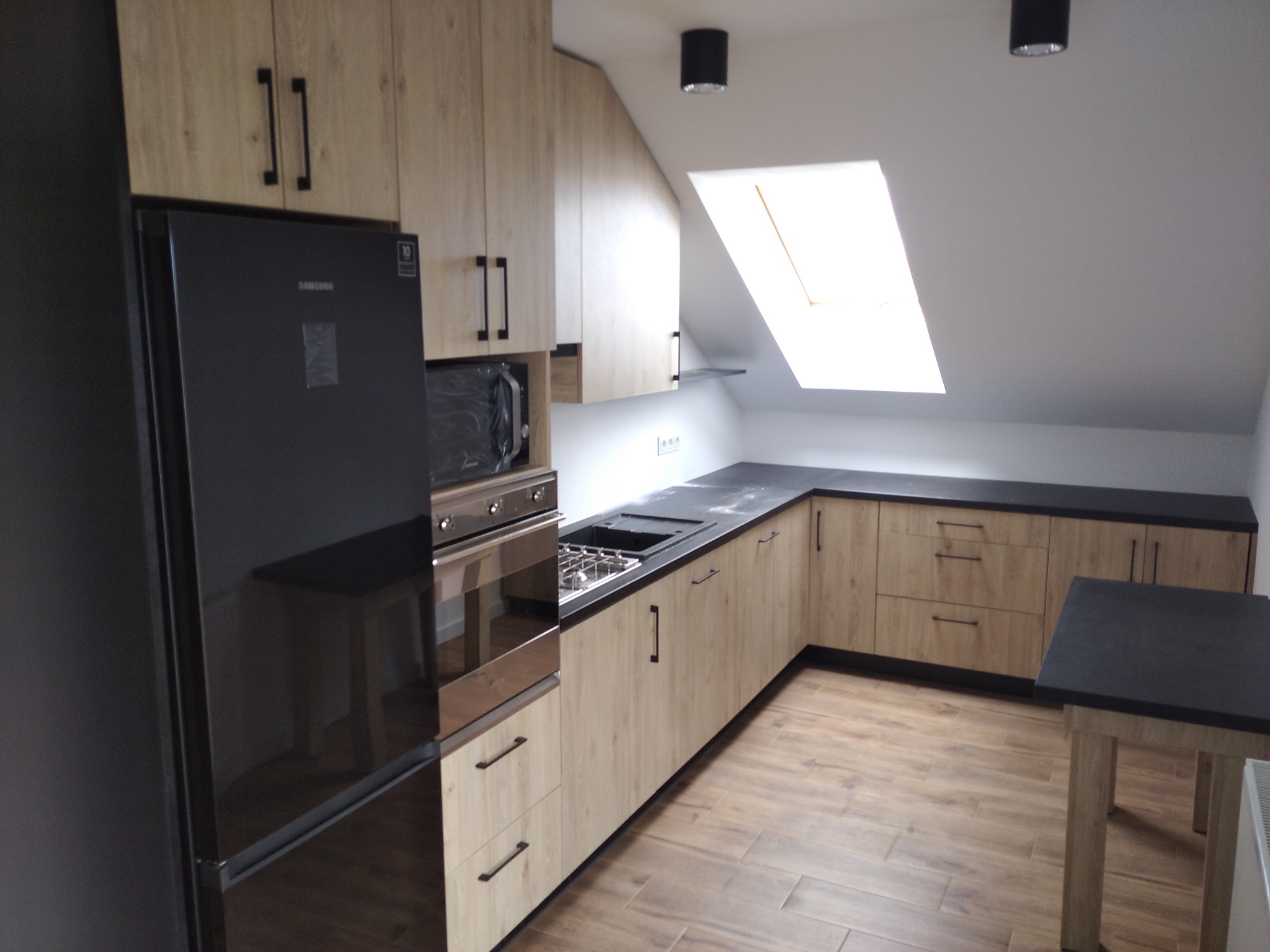 Jasna kuchnia w skandynawskim stylu na poddaszu ze skośnym sufitem i oknem dachowym, czarne blaty i sprzęt AGD, drewniana podłoga.