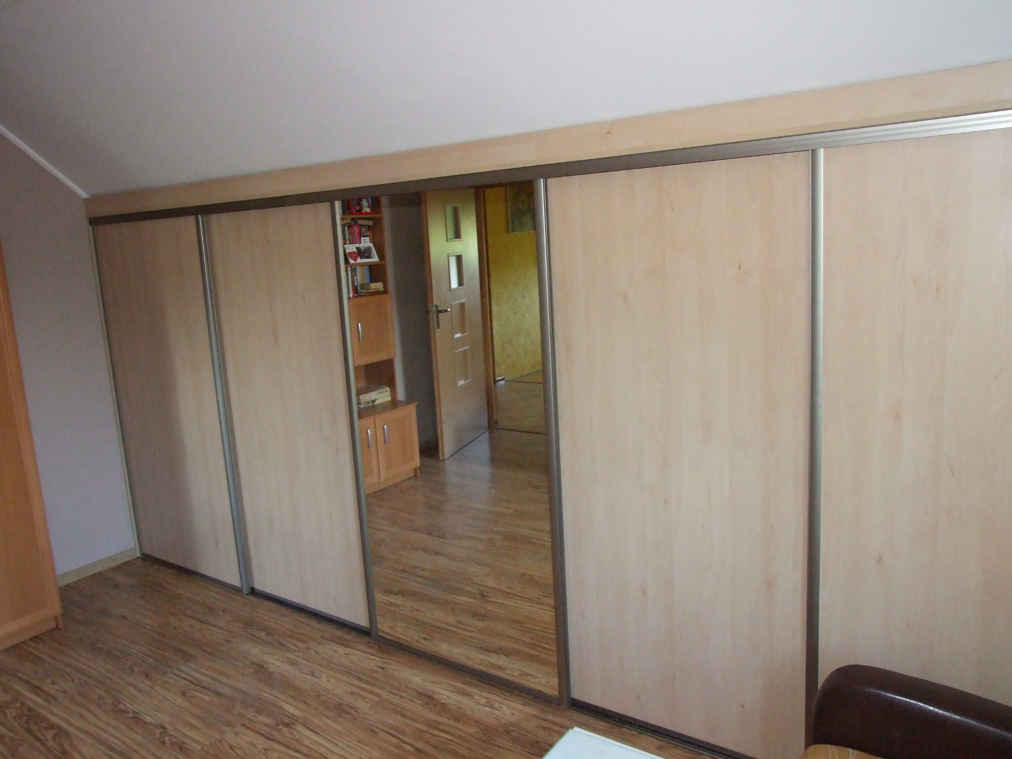 Duża szafa wnękowa z przesuwnymi drzwiami z jasnego drewna i lustrzanym panelem, odbijającym fragment pokoju z drewnianą podłogą i regałem.