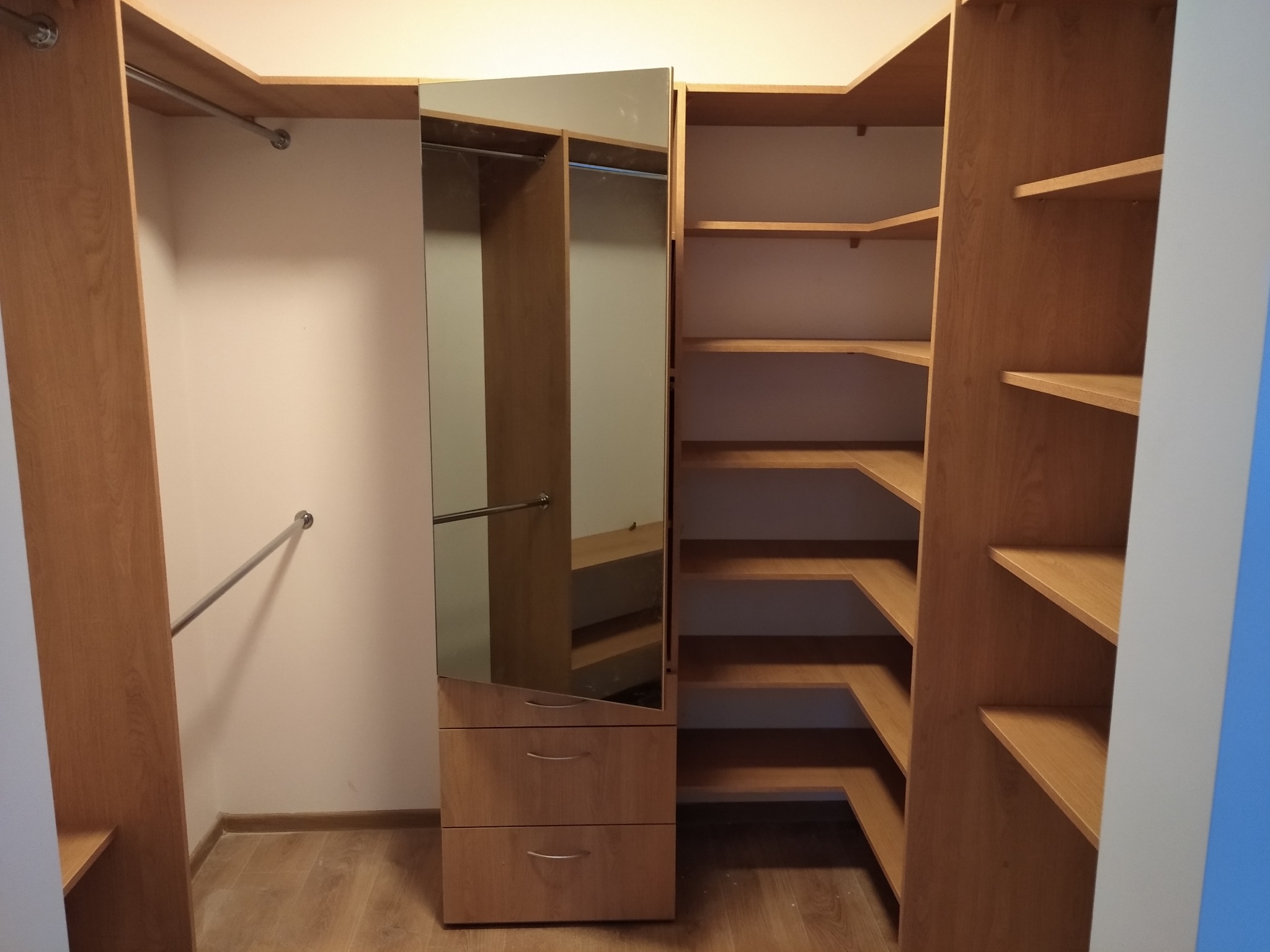 Jasna, drewniana garderoba z lustrzanymi drzwiami i półkami narożnymi, widok na wnętrze z drążkami na wieszaki i trzema szufladami.