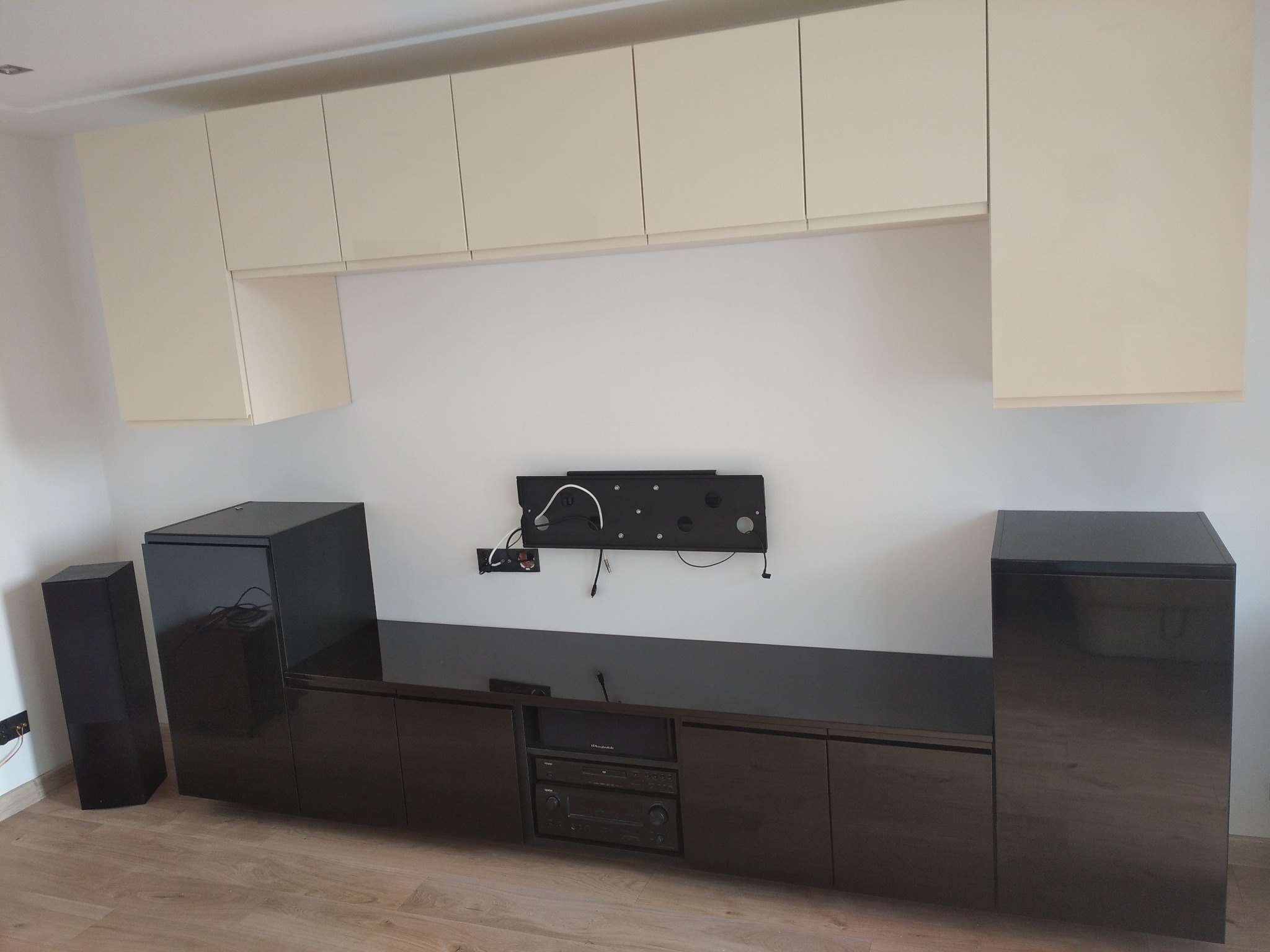 Jasne szafki wiszące nad ciemnobrązową zabudową RTV z kolumnami głośnikowymi po bokach, zamontowane na ścianie uchwyty pod telewizor.