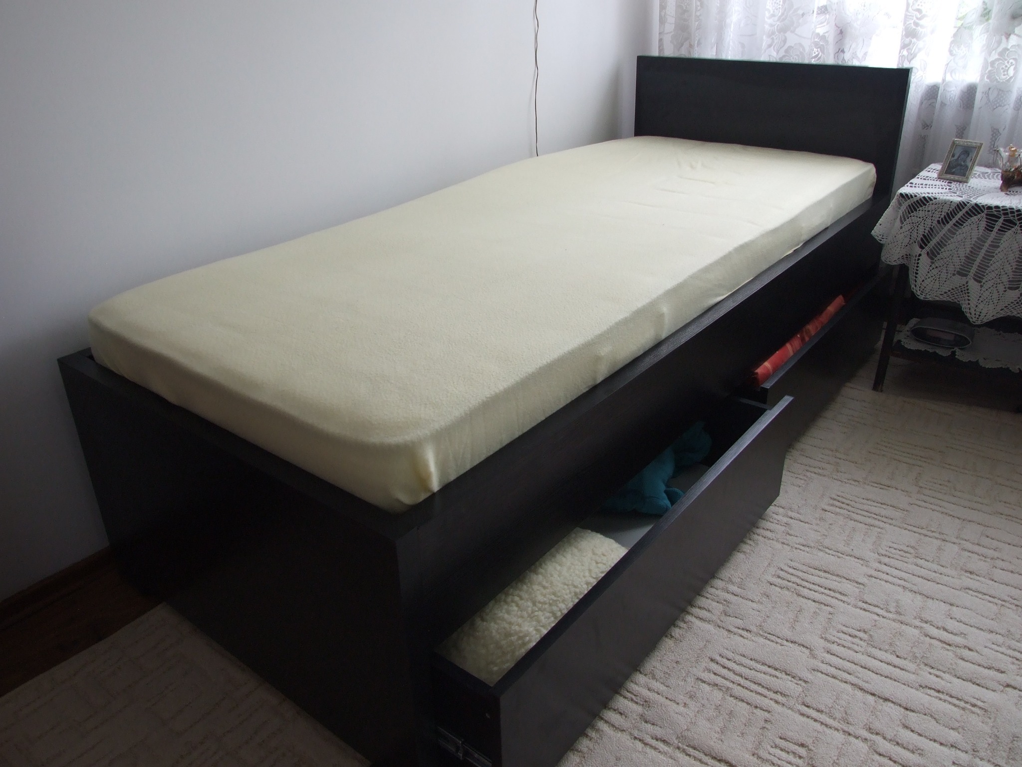 Łóżko jednoosobowe z ciemnego drewna z wysuwaną szufladą, w której widać zwinięty jasny koc i niebieską maskotkę, na beżowym dywanie.