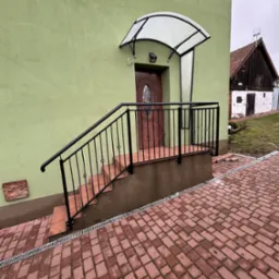 Zewnętrzne schody wejściowe z czarną, metalową balustradą z ozdobnymi elementami, prowadzące do drzwi wejściowych z zadaszeniem z poliwęglanu, na tle zielonej elewacji budynku i kostki brukowej.