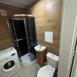 Ukończona łazienka z kabiną prysznicową, umywalką z szafką, toaletą i pralką, wykończona płytkami imitującymi drewno na ścianach.