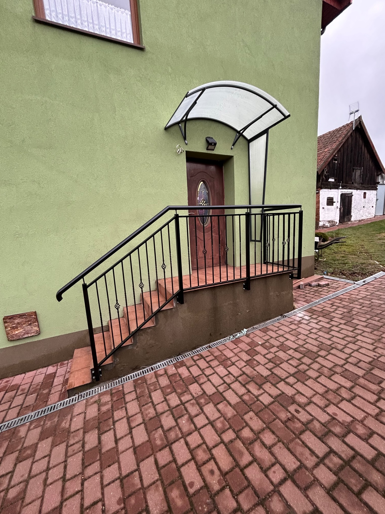 Zewnętrzne schody wejściowe z czarną, metalową balustradą z ozdobnymi elementami, prowadzące do drzwi wejściowych z zadaszeniem z poliwęglanu, na tle zielonej elewacji budynku i kostki brukowej.