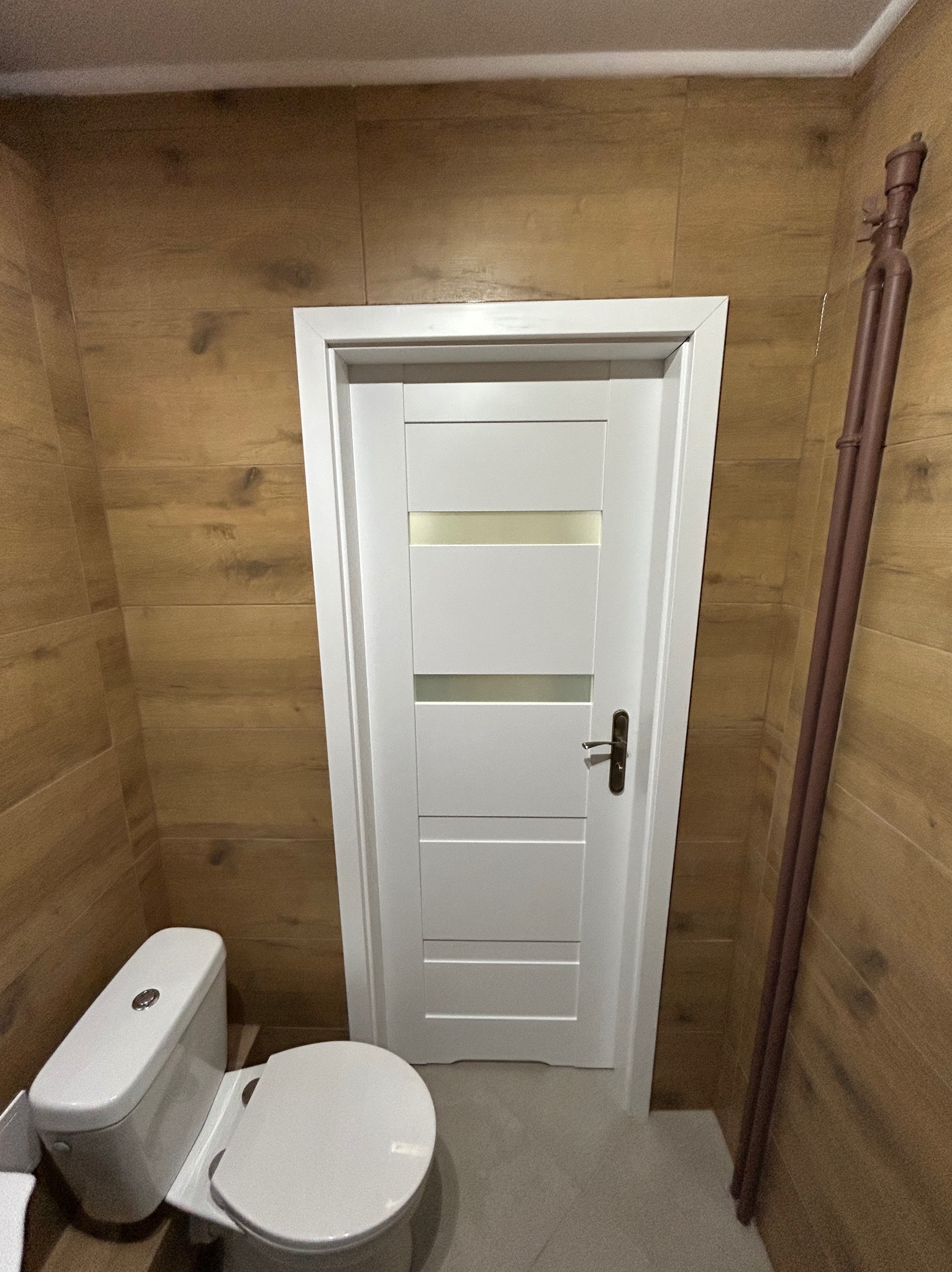 Wykończona łazienka w Biskupcu z toaletą i białą, nowoczesną stolarką drzwiową na tle drewnopodobnych płytek ściennych oraz brązowych rur grzewczych.