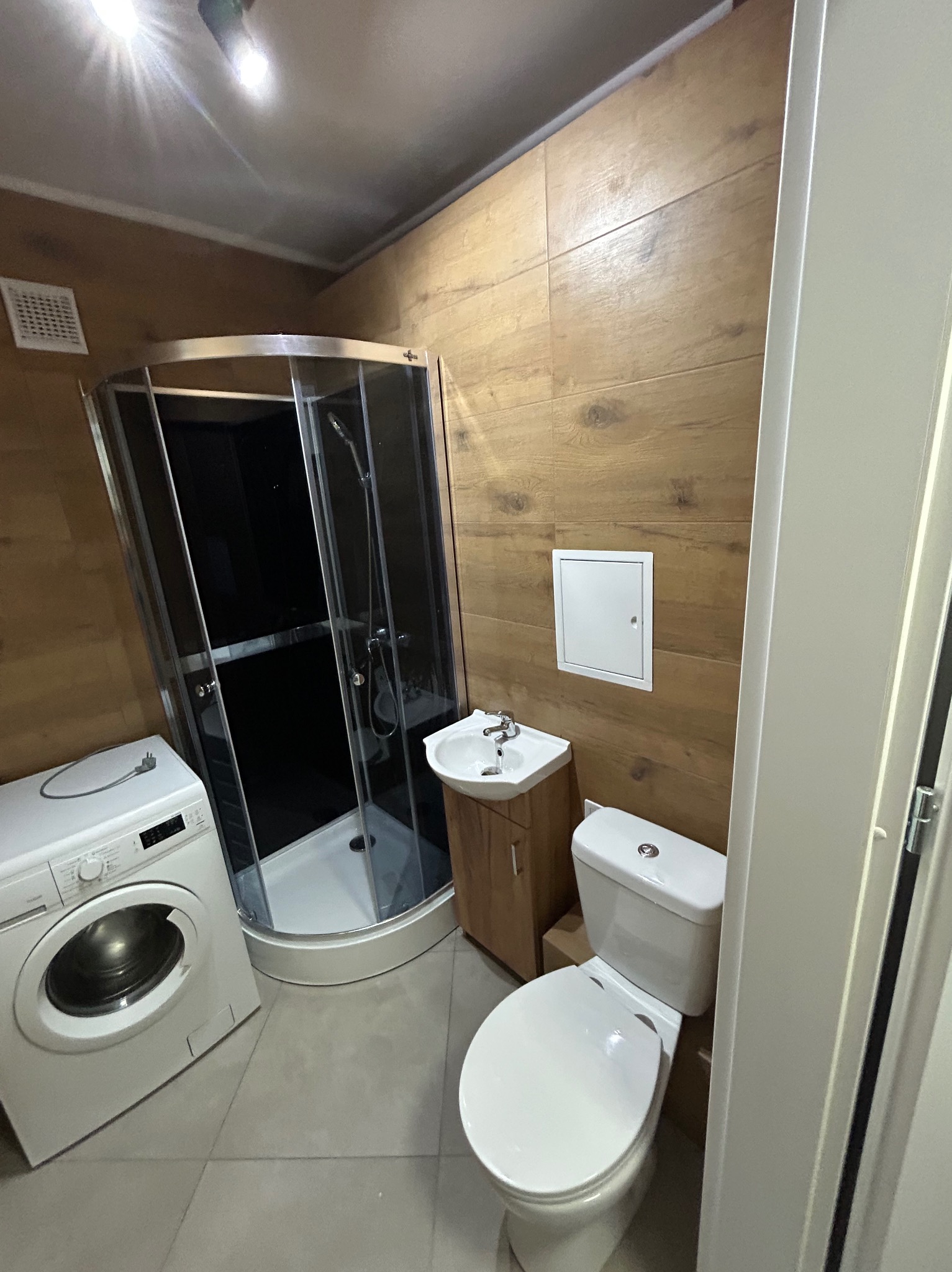 Ukończona łazienka z kabiną prysznicową, umywalką z szafką, toaletą i pralką, wykończona płytkami imitującymi drewno na ścianach.