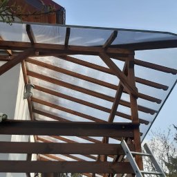 Usługi Dekarsko - Ciesielskie Grzegorz Lubowiecki - Drewniana konstrukcja z przezroczystym zadaszeniem z poliwęglanu, widok z dołu, drabina oparta o konstrukcję, błękitne niebo w tle.