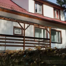 Dwupiętrowy dom z czerwonym dachem z dachówki, drewnianymi obróbkami okiennymi i drewnianą konstrukcją balkonu, widziany znad porośniętego bluszczem ogrodzenia.