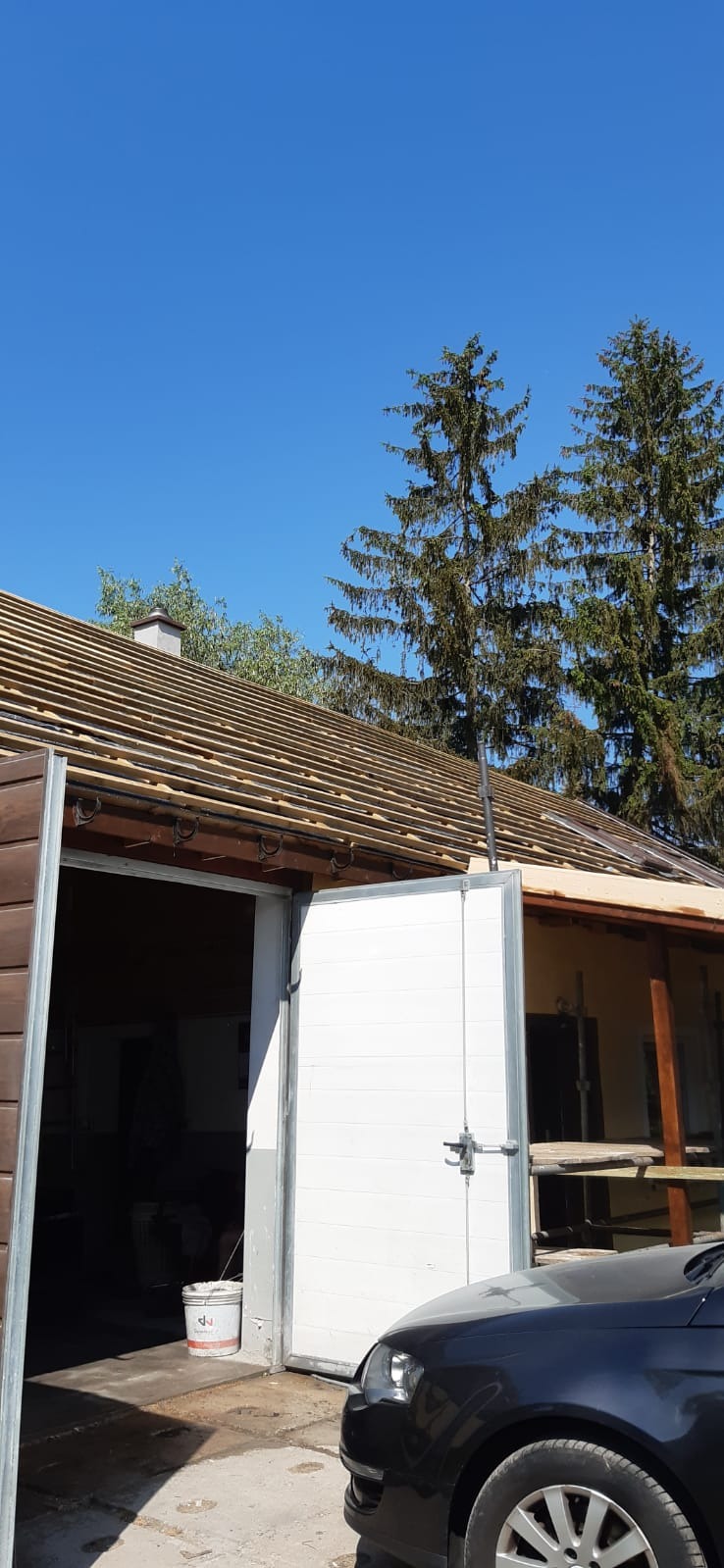 Wstępny etap prac nad dachem z widoczną konstrukcją łat i podłatników. W tle wysokie drzewa iglaste i błękitne niebo. Częściowo widoczny budynek z otwartą białą bramą garażową i zaparkowany samochód.