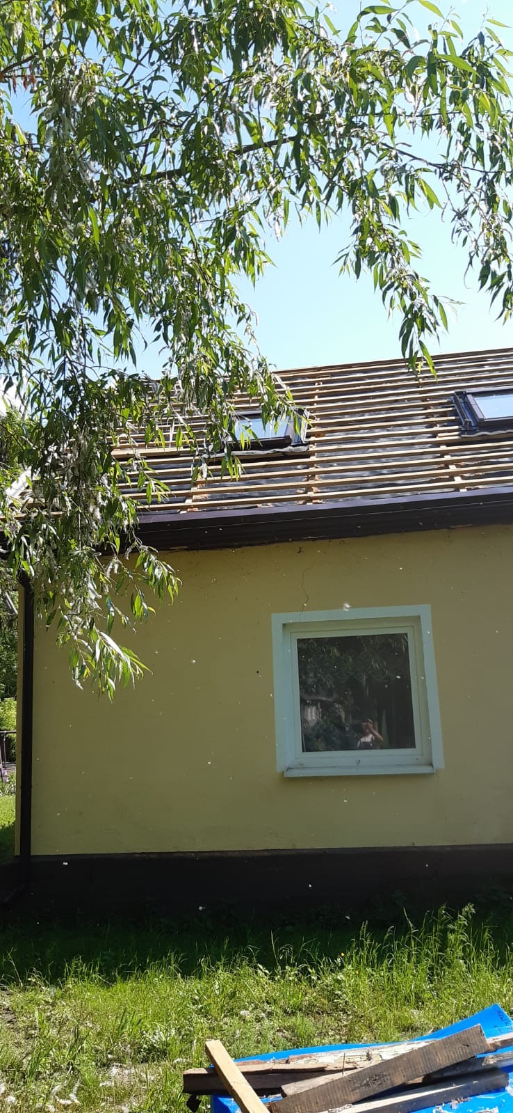 Dom jednorodzinny z dachem w trakcie wymiany, widoczne łaty pod nową dachówkę i okna dachowe, częściowo przysłonięty gałęziami drzewa