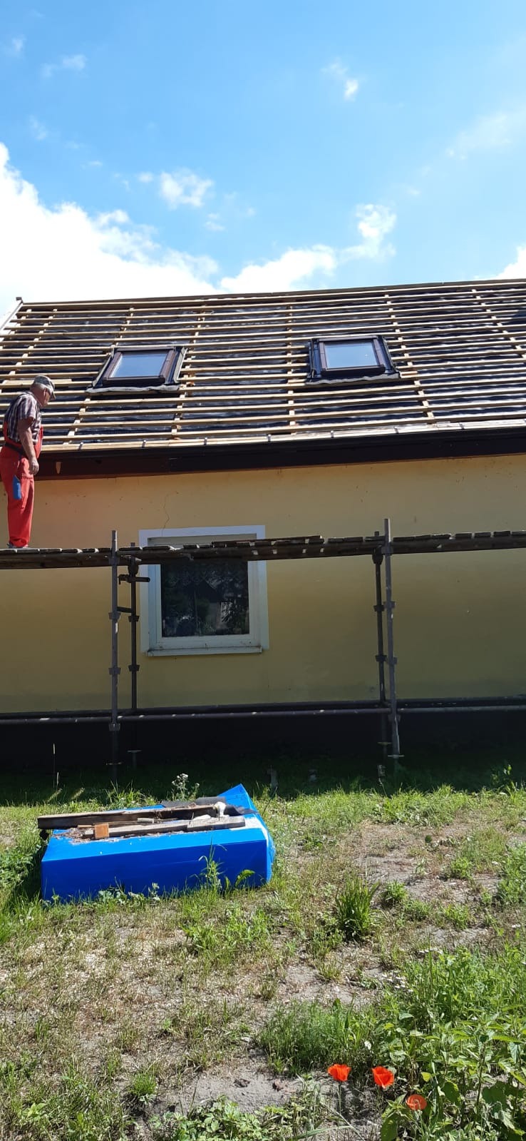 Pracownik w czerwonym kombinezonie na rusztowaniu układa łatki pod dachówki wokół okien dachowych na tle błękitnego nieba z chmurami.