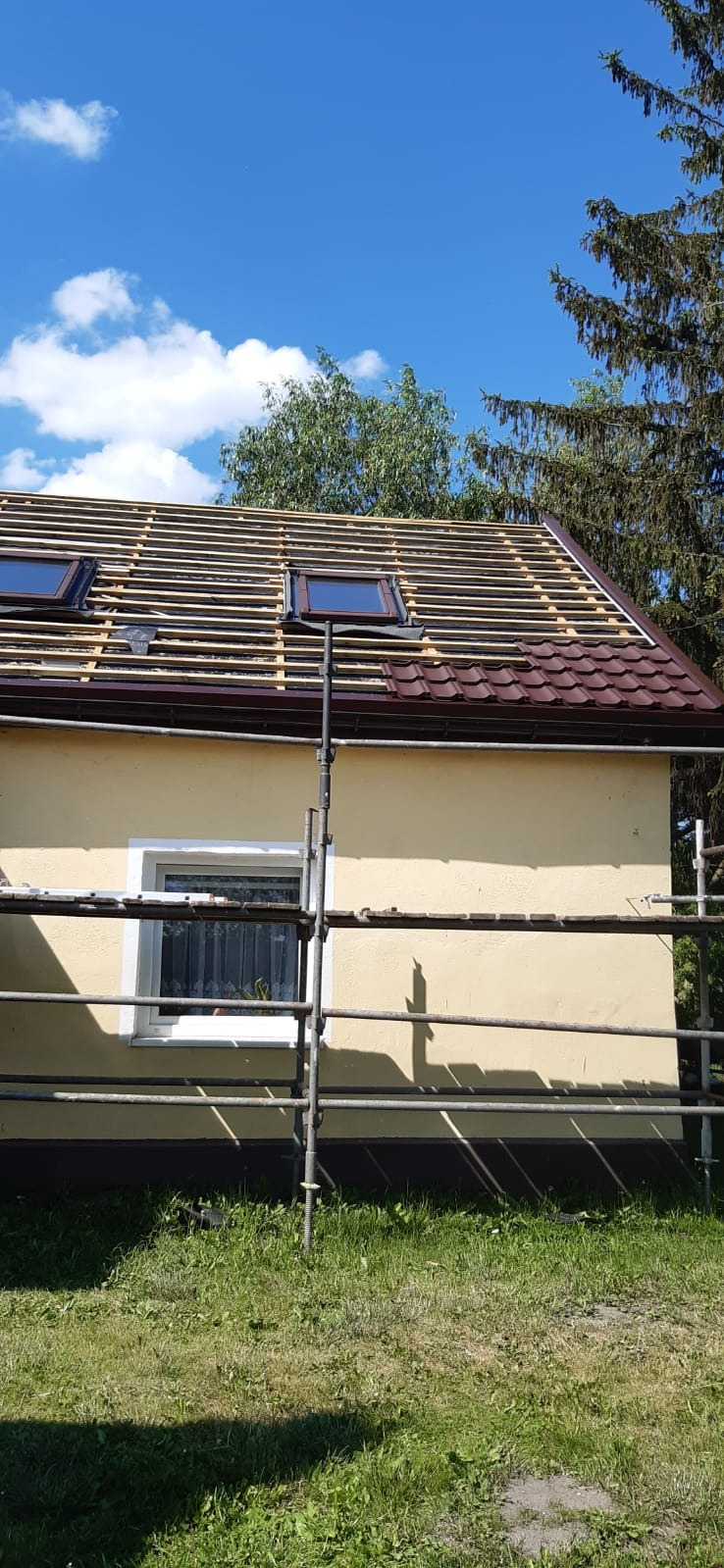 Dom w trakcie krycia dachu: widoczne łaty, dwa okna dachowe i częściowo ułożona brązowa dachówka. Ustawione rusztowanie przy ścianie budynku.