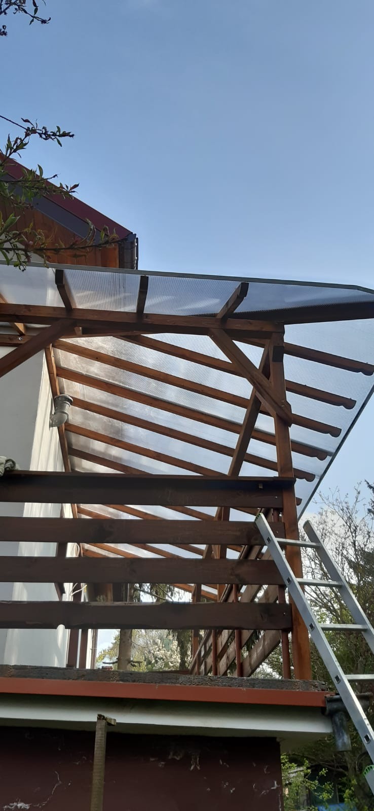 Drewniana konstrukcja z przezroczystym zadaszeniem z poliwęglanu, widok z dołu, drabina oparta o konstrukcję, błękitne niebo w tle.