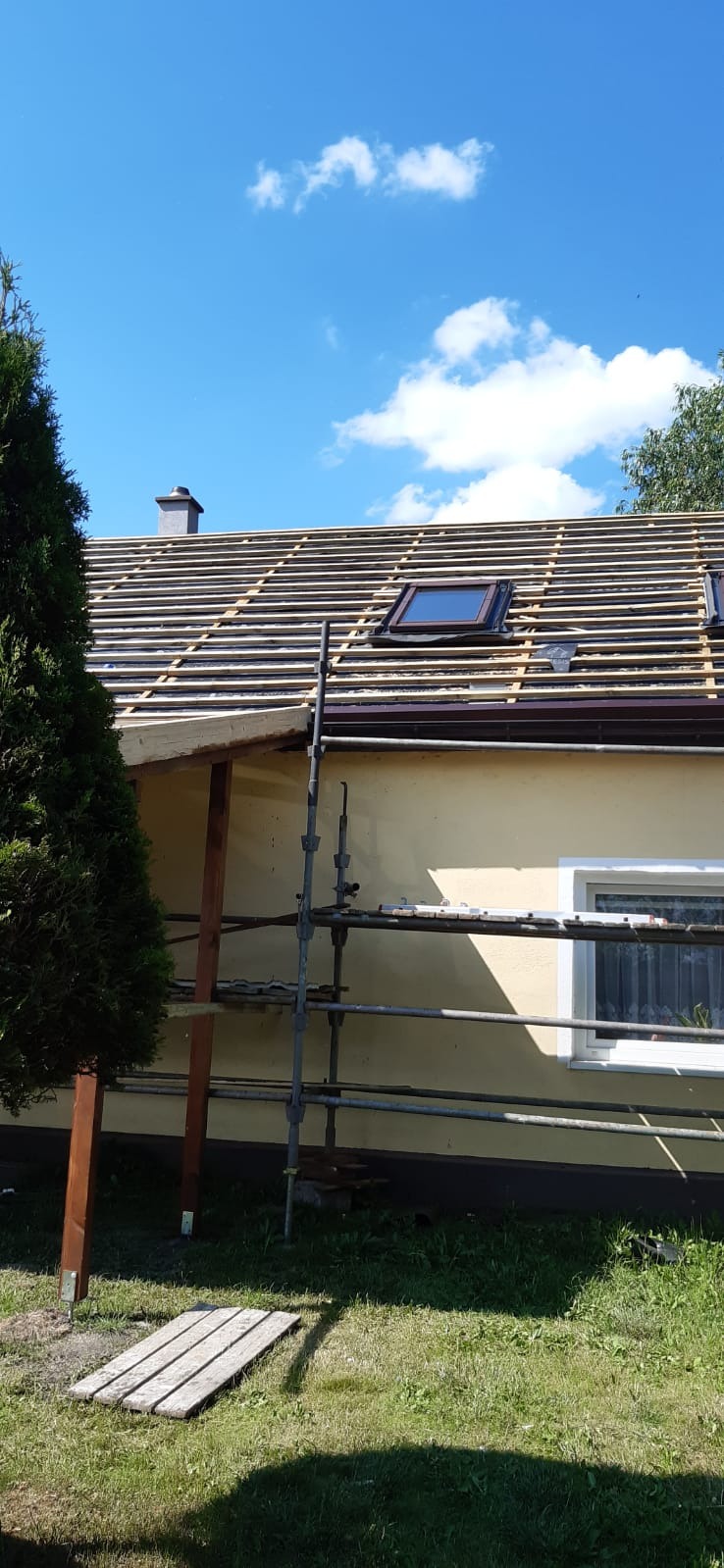 Dom jednorodzinny w trakcie przygotowania do położenia nowego pokrycia dachowego, widoczne łaty, okno dachowe i komin na tle błękitnego nieba z chmurami.