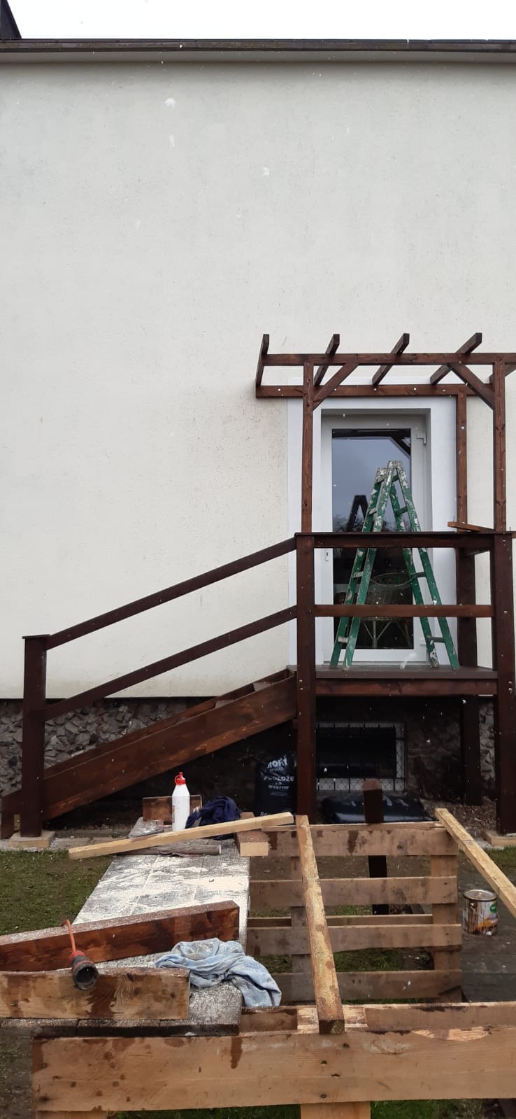 Drewniana konstrukcja ganku z balustradą i zadaszeniem nad drzwiami, w trakcie budowy, widoczna drabina oparta o drzwi, rozłożone deski, narzędzia i materiały budowlane.