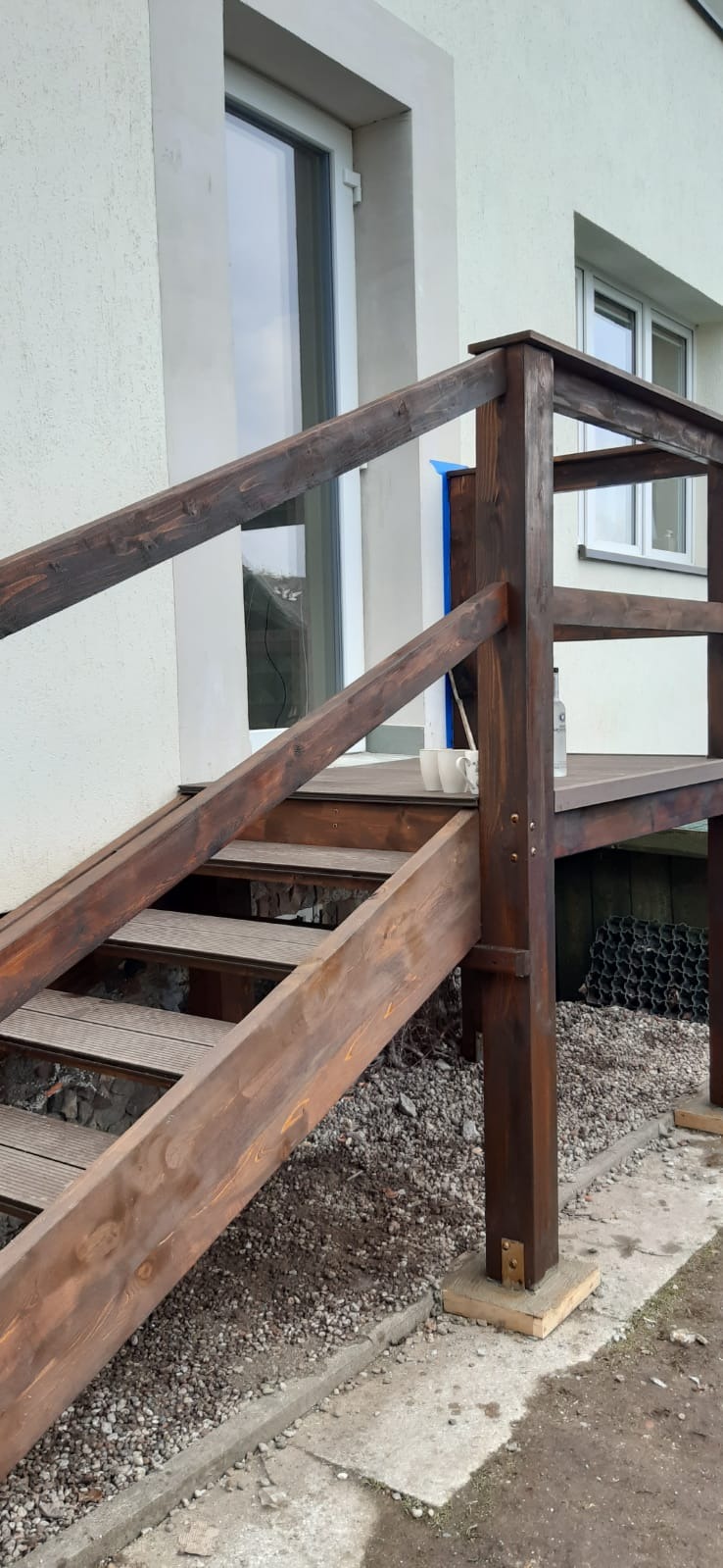 Drewniane schody wejściowe z balustradą, świeżo pomalowane bejcą w kolorze ciemnego brązu, prowadzące do białych drzwi wejściowych domu.