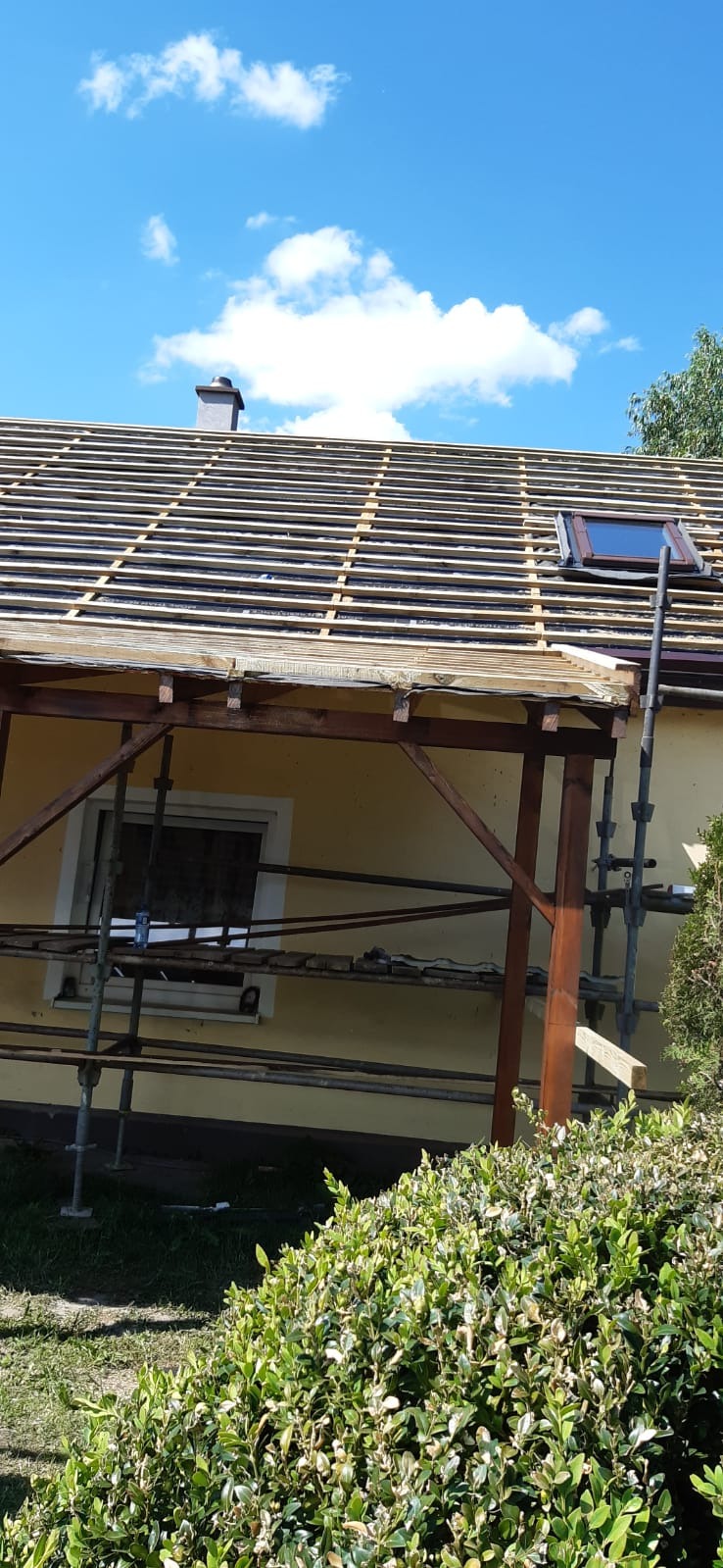 Przygotowanie podłoża pod nowe pokrycie dachowe z widocznym oknem dachowym i kominem na tle błękitnego nieba z chmurami, rusztowanie przy elewacji budynku.