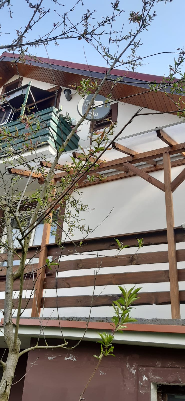 Dom z drewnianą pergolą i balkonem z zieloną balustradą, widok z dołu przez gałęzie drzewa.