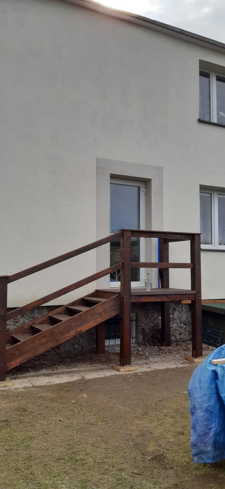 Drewniane schody wejściowe z balustradą, prowadzące do drzwi budynku z jasną elewacją, widoczne niedoskonałości w wykończeniu terenu.