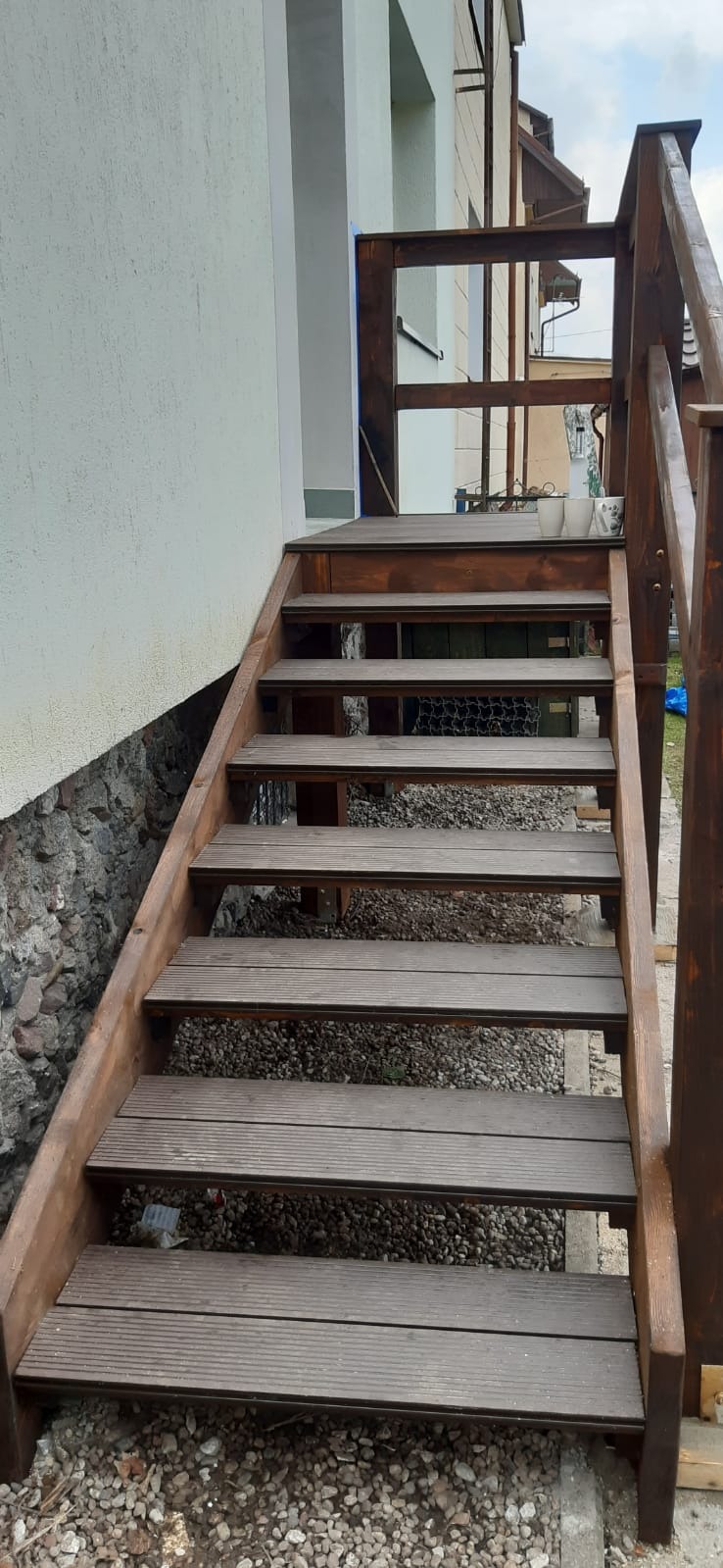 Drewniane schody zewnętrzne prowadzące do budynku, z widoczną konstrukcją nośną i deskami z ryflowanej powierzchni, obok ściana budynku z kamienną podmurówką.