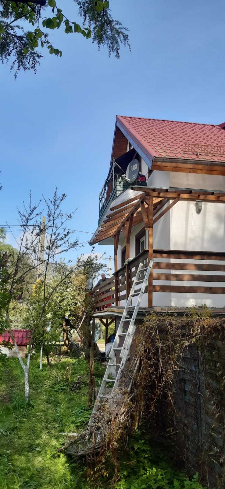 Dom z czerwonym dachem widziany z ogrodu, z widoczną drabiną opartą o ścianę i elementami drewnianej konstrukcji werandy.