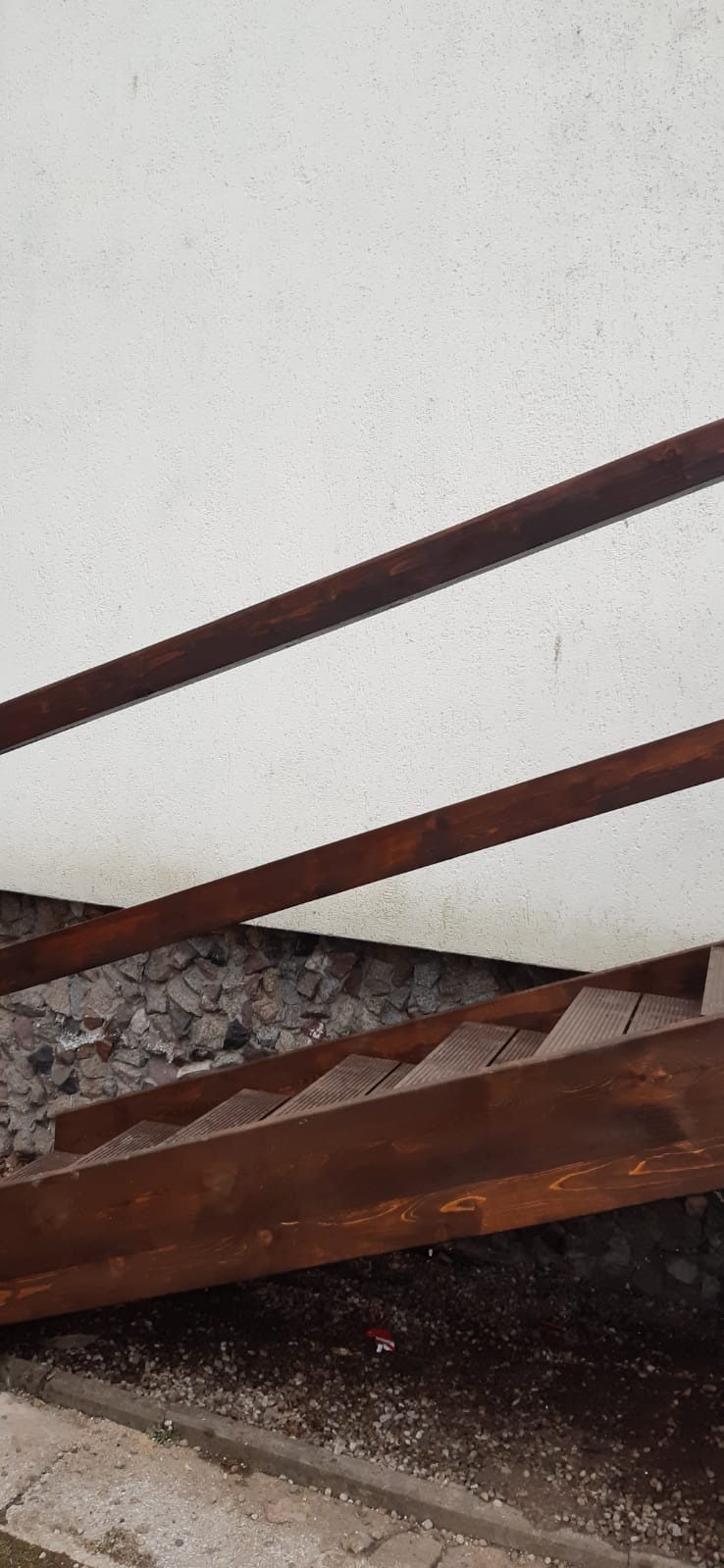 Drewniane schody zewnętrzne z brązową balustradą prowadzące w dół, z widoczną ścianą wykończoną kamieniem i fragmentem białej ściany powyżej.