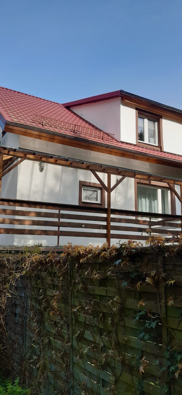 Dom z czerwonym dachem krytym dachówką, drewnianą pergolą i ogrodzeniem porośniętym bluszczem w jesiennej scenerii.