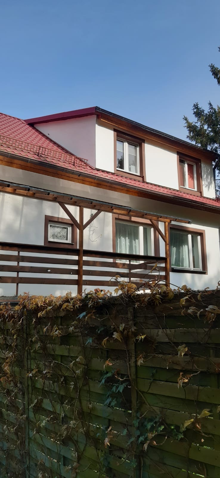 Dwupiętrowy dom z czerwonym dachem z dachówki, drewnianymi obróbkami okiennymi i drewnianą konstrukcją balkonu, widziany znad porośniętego bluszczem ogrodzenia.