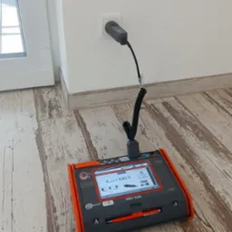 Miernik Sonel MPI-540 podłączony do gniazdka elektrycznego, wyświetlający wartość impedancji pętli zwarcia ZL-PE = 0,82 Ω, na podłodze z paneli imitujących drewno.