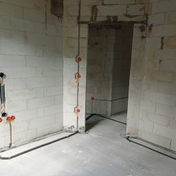 Skrobak Jan - Wnętrze surowego budynku z widoczną instalacją elektryczną w peszlu i puszkami podtynkowymi na ścianach z betonu komórkowego, napis 'ZASILANA' na ścianie.