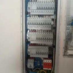 Rozdzielnica elektryczna z zainstalowanymi bezpiecznikami, wyłącznikami różnicowoprądowymi i ogranicznikami przepięć, widoczne okablowanie w kolorach niebieskim, żółto-zielonym i brązowym.