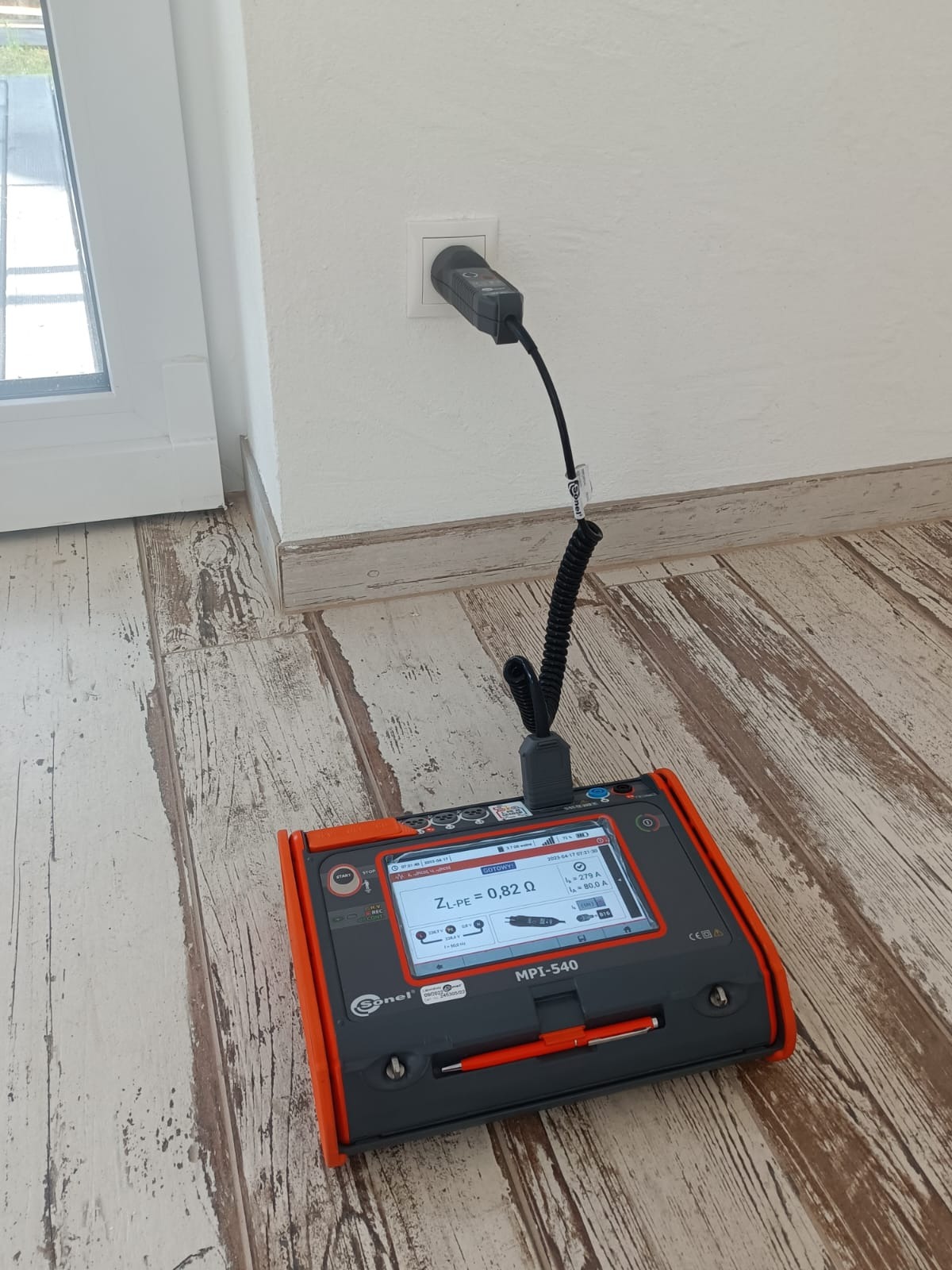Miernik Sonel MPI-540 podłączony do gniazdka elektrycznego, wyświetlający wartość impedancji pętli zwarcia ZL-PE = 0,82 Ω, na podłodze z paneli imitujących drewno.