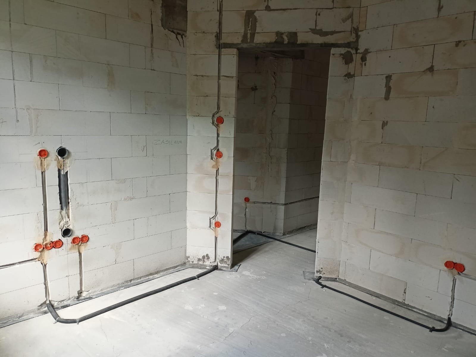 Wnętrze surowego budynku z widoczną instalacją elektryczną w peszlu i puszkami podtynkowymi na ścianach z betonu komórkowego, napis 'ZASILANA' na ścianie.
