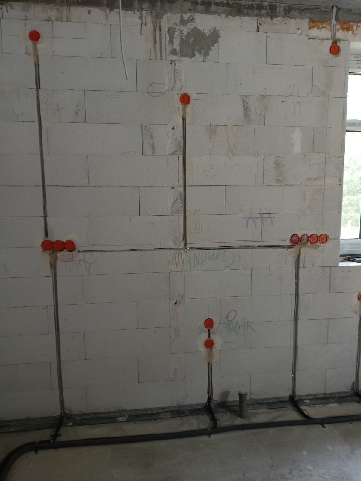Surowa ściana z betonu komórkowego z widocznymi przewodami elektrycznymi w peszlach i puszkami instalacyjnymi, przygotowana do dalszych prac wykończeniowych.