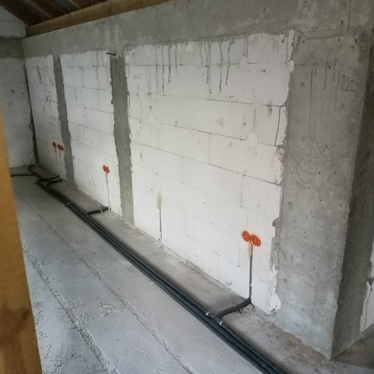 Wnętrze w trakcie prac elektrycznych: kable w peszlach ułożone wzdłuż ściany z betonu komórkowego, widoczne puszki elektryczne w kolorze pomarańczowym, surowy stan wykończenia.