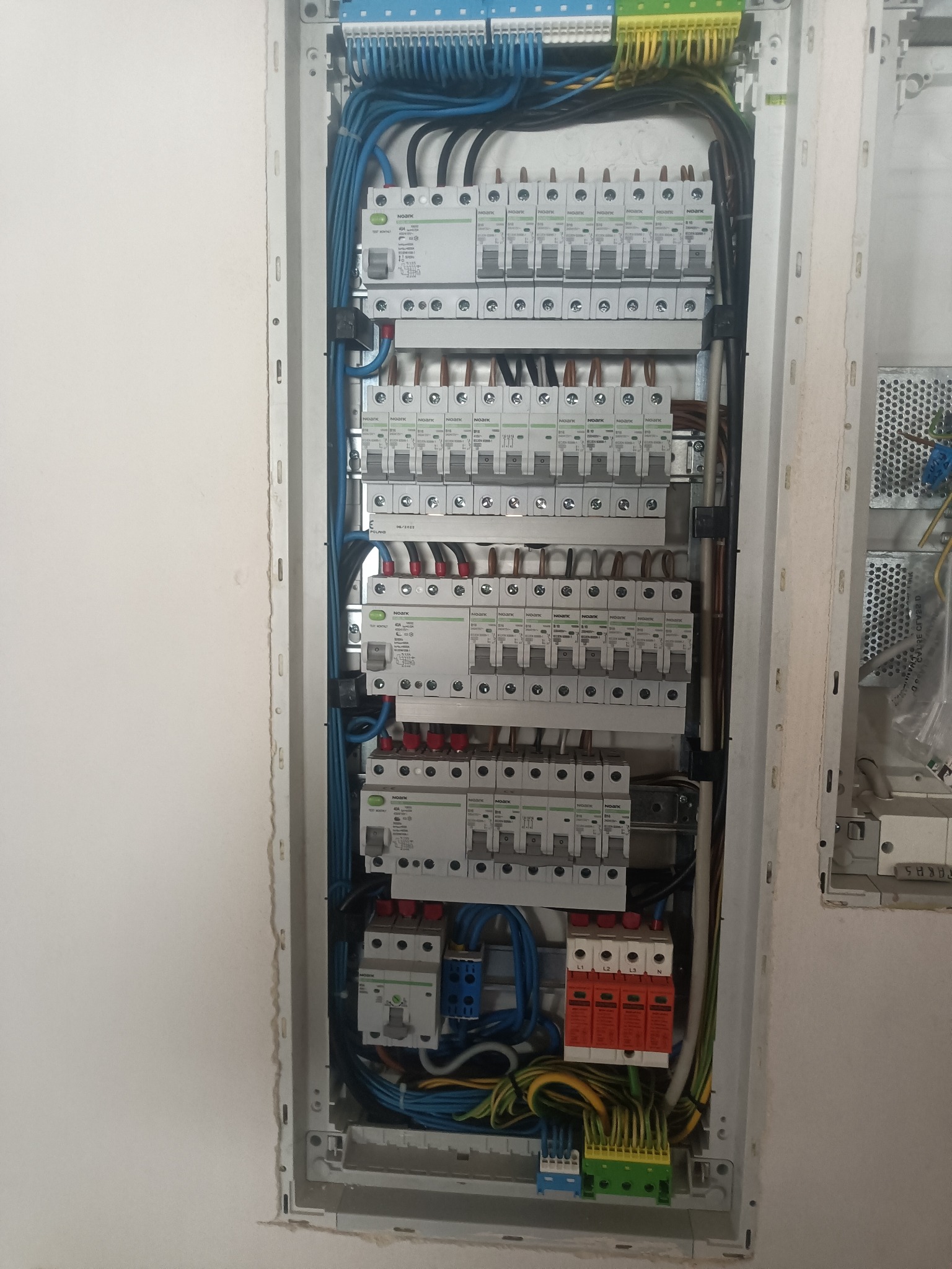 Rozdzielnica elektryczna z zainstalowanymi bezpiecznikami, wyłącznikami różnicowoprądowymi i ogranicznikami przepięć, widoczne okablowanie w kolorach niebieskim, żółto-zielonym i brązowym.