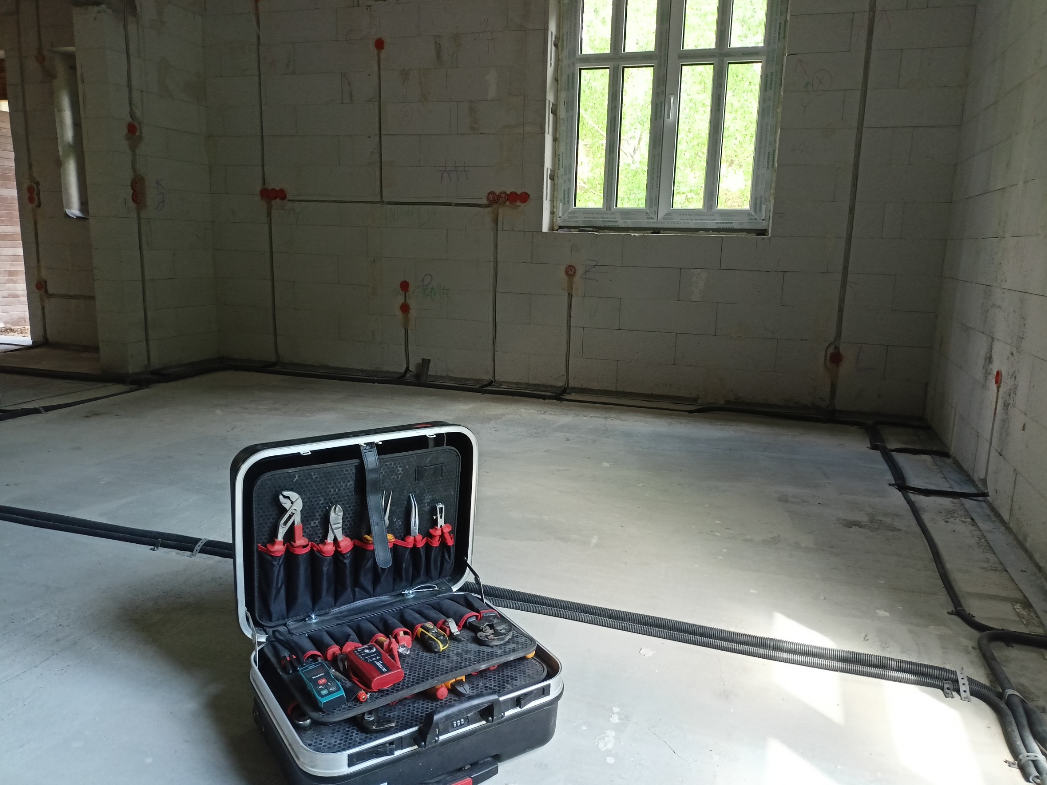 Wnętrze surowego pomieszczenia z widocznymi przewodami elektrycznymi w peszlach, zamontowanymi na ścianach z betonu komórkowego, oraz otwartą skrzynką narzędziową elektryka na pierwszym planie.